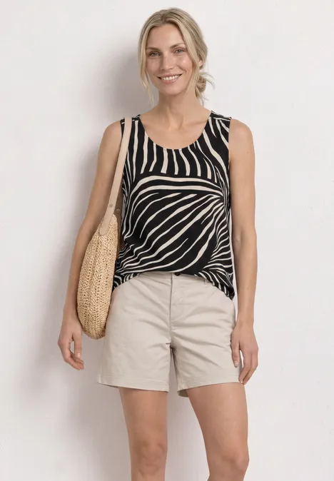 Frau in Zebra-Print Top, beigen Shorts und Strohtasche vor weißer Wand.