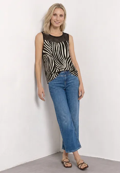 Eine junge blondhaarige Frau lehnt vor einer weißen Wand, trägt ein Zebra-Top, gerade Jeans und Sandalen.