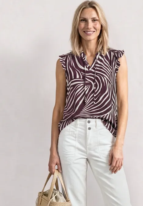 Top mit Strukturmix und Crochet-Details rustic rouge Eine Frau trägt eine ärmellose Bluse im Zebramuster, weiße Hose und hält eine Strohtasche.