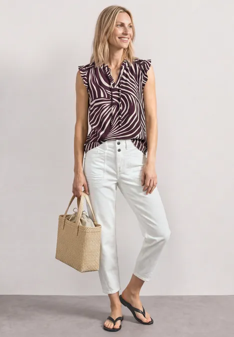 Top mit Strukturmix und Crochet-Details rustic rouge Frau trägt zebra-muster Top, weiße Jeans, schwarze Flip-Flops und hält eine Korbtasche vor neutralem Hintergrund.