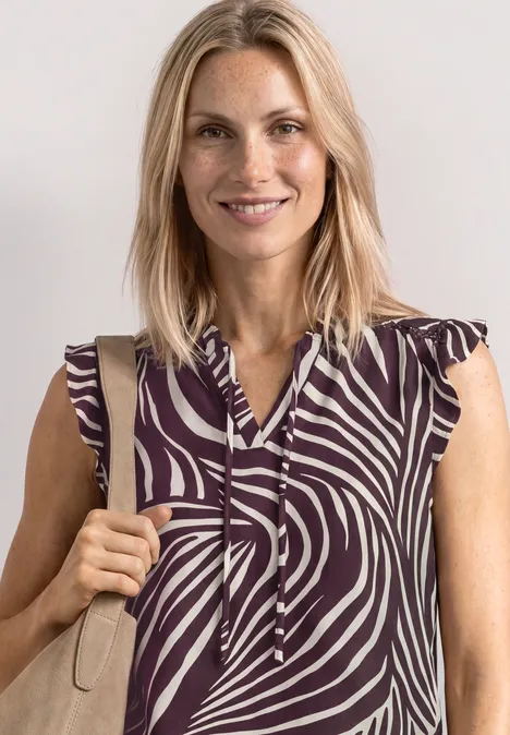 Top mit Strukturmix und Crochet-Details rustic rouge Blonde Frau trägt ärmelloses Top mit lila-beigem Zebramuster und hält einen beigen Taschenriemen.