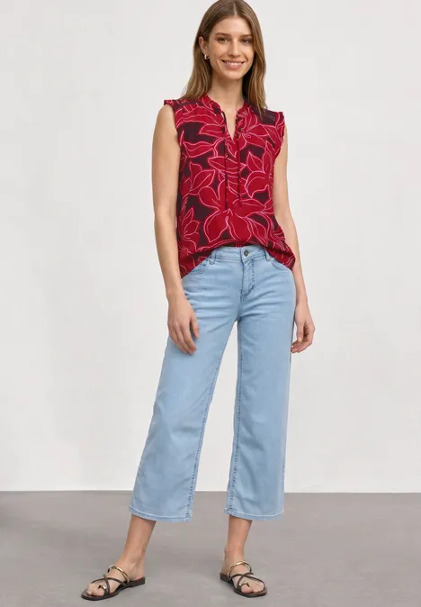 Model trägt rote ärmellose Bluse mit Blumenmuster, hellblaue Culotte-Jeans und schwarze Sandalen vor weißem Hintergrund.