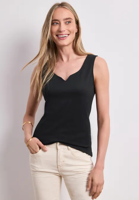 Vrouw in zwarte tanktop en lichte broek, staand voor een neutrale achtergrond.