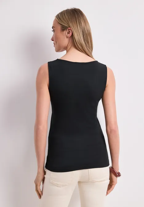 Een vrouw van achteren voor een neutrale achtergrond, draagt een zwart mouwloos topje en een lichte broek.