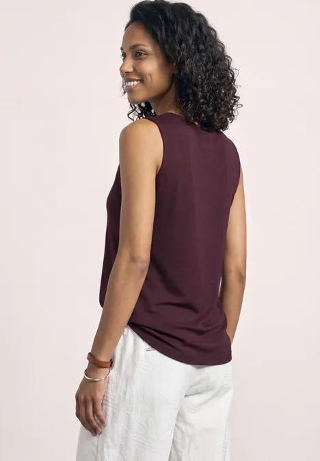Basic top met hartvormige halslijn rustic rouge Person mit lockigem Haar und unscharfem Gesicht von hinten, in ärmellosem bordeauxfarbenem Top und weißen Hosen.