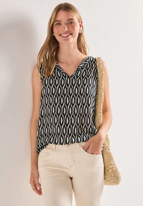 Vrouw in mouwloze blouse met patroon, beige broek en strozak over de schouder, hand in de zak.