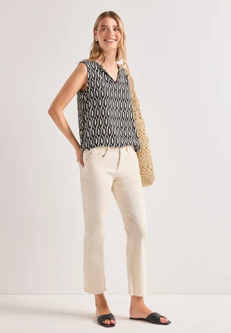 Vrouw met mouwloze top met print, lichte broek, zwarte sandalen en een strooien tas.