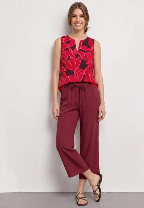 Vrouw staat voor witte muur in rood topje met bladmotief, wijde bordeauxrode broek en zwarte sandaaltjes.