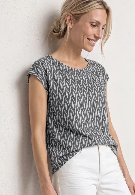 Femme appuyée au mur, porte un top noir et blanc à manches courtes et un pantalon blanc.