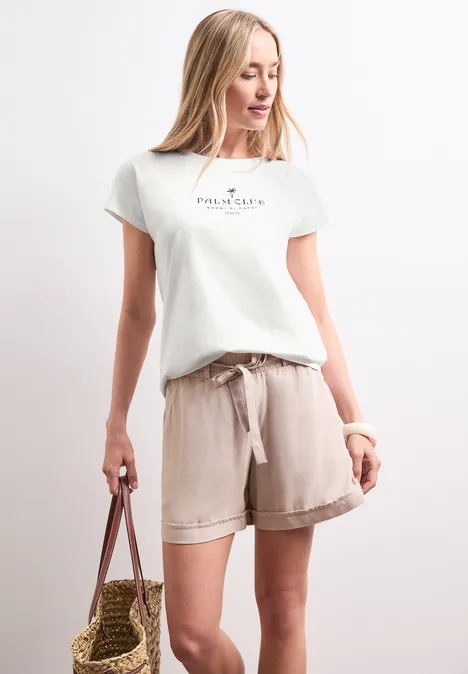 Koszulka na ramiączkach z napisem off white Kobieta ubrana w biały t-shirt z nadrukiem i beżowe lniane szorty, trzyma słomkową torbę na białym tle.