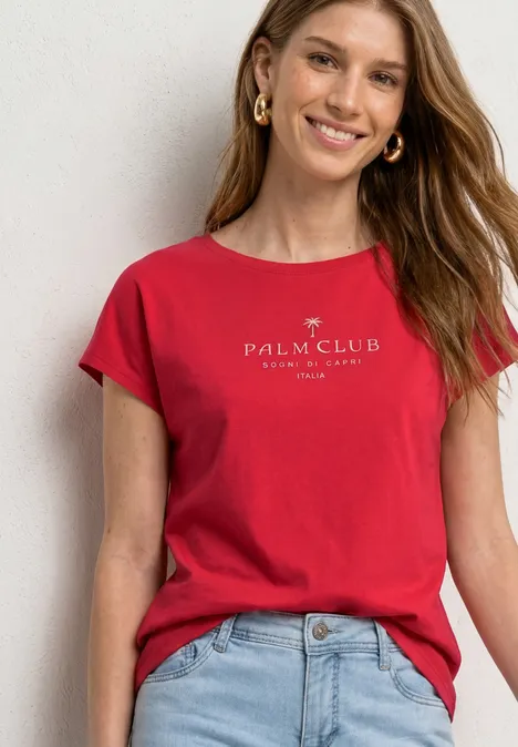 Dropped Shoulder Shirt mit Wording salsa red Frau trägt rotes T-Shirt mit Palm Club Logo und hellblauer Jeans vor weißer Wand.