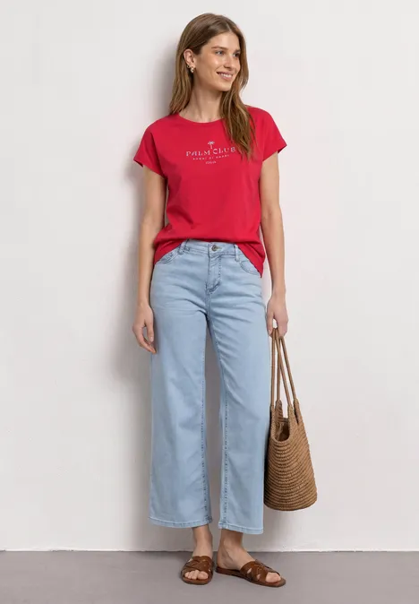 Dropped Shoulder Shirt mit Wording salsa red Eine Frau in rotem T-Shirt, hellblauen Jeans, Sandalen und Korbtasche vor weißer Wand.