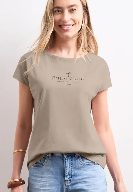 Koszulka na ramiączkach z napisem clay sand Kobieta ma na sobie beżowy t-shirt z napisem 'Palm & Claw Culture' i niebieskie dżinsy.