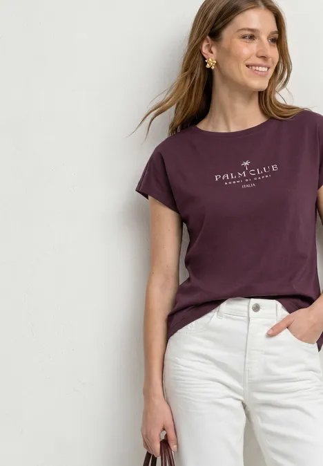 Dropped Shoulder Shirt mit Wording rustic rouge Frau in weinrotem Paul & Shark T-Shirt und weißen Jeans lehnt an weißer Wand.