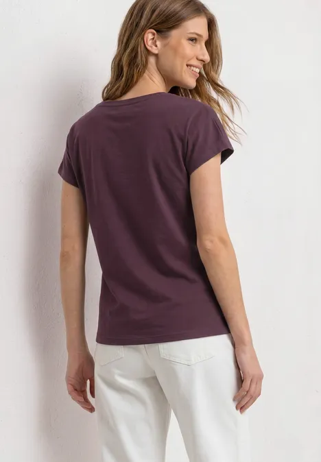Dropped Shoulder Shirt mit Wording rustic rouge Frau steht mit dem Rücken zur Kamera, trägt violettes T-Shirt und weiße Hose vor hellem Hintergrund.