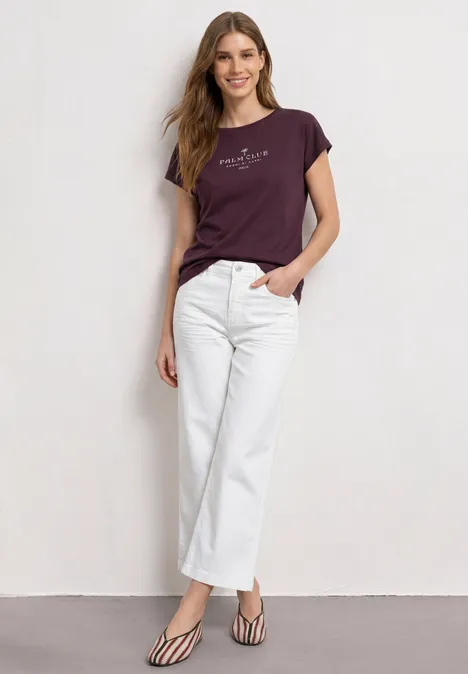 Dropped Shoulder Shirt mit Wording rustic rouge Eine Frau steht vor einer Wand in weißer Hose, lila T-Shirt und gestreiften High-Heels.