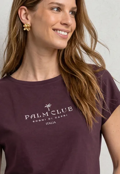 Dropped Shoulder Shirt mit Wording rustic rouge Frau mit welligem Haar, goldenen Blüten-Ohrringen und dunkelviolettem T-Shirt mit 'Palm Club'-Schriftzug