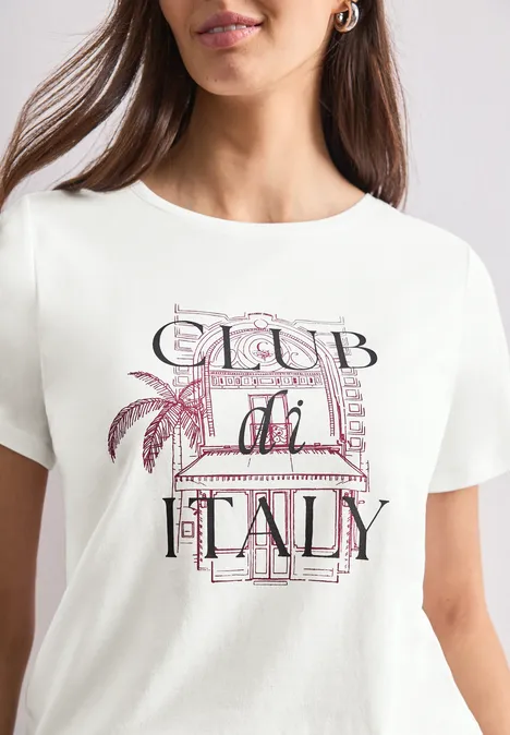 Frau trägt weißes T-Shirt mit 'Club di Italy'-Aufdruck, pastellfarbene Buchstaben und Palmenmotiv.