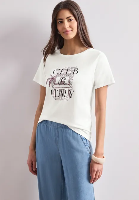 Eine Frau trägt ein weißes T-Shirt mit Aufdruck und lockere, hellblaue Hose vor neutralem Hintergrund.