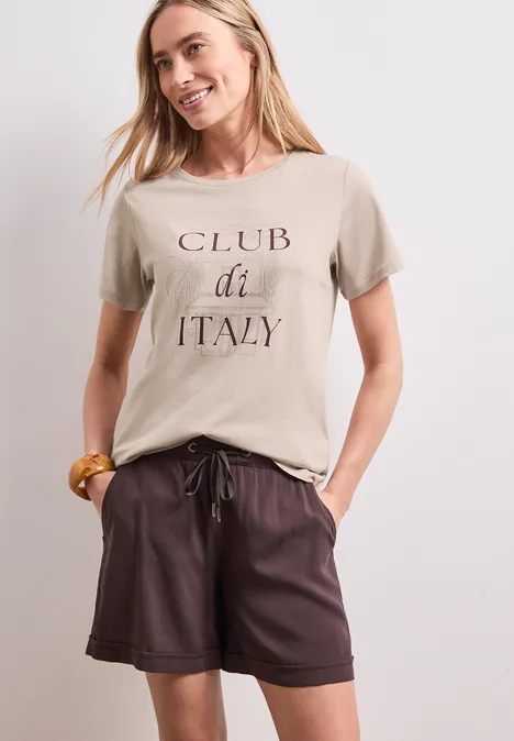 Frau trägt beiges T-Shirt mit 'CLUB di ITALY'-Aufdruck und braune Leder-Shorts.