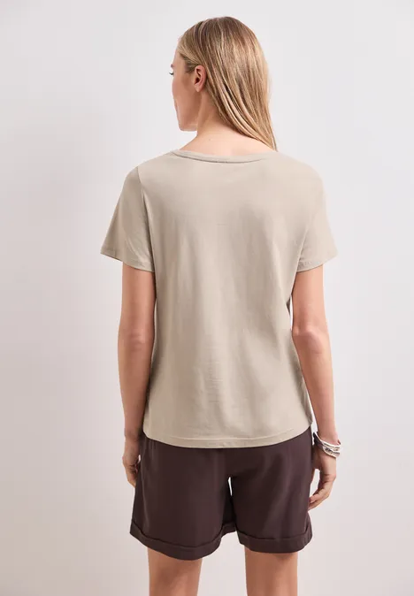 Rückansicht einer blonden Frau in beige T-Shirt und braunen Shorts vor hellem Hintergrund.