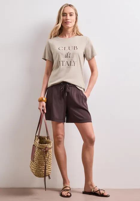 Frau in legerer Sommerkleidung: T-Shirt, Shorts, Sandalen, hält eine Strohtasche.