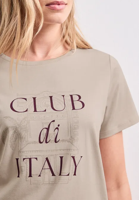 Eine Frau trägt ein beiges T-Shirt mit dem Aufdruck "CLUB di ITALY" und dezenten Architekturzeichnungen.