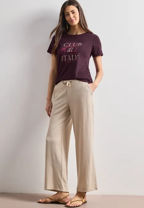 Eine Frau steht in minimalistischem Umfeld, trägt ein lila T-Shirt mit 'Club de Italy', beige weite Hose und Sandalen.
