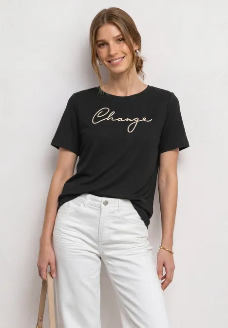 Frau trägt schwarzen T-Shirt mit 'Change'-Schriftzug, weiße Hose, hält Tasche, vor hellem Hintergrund.