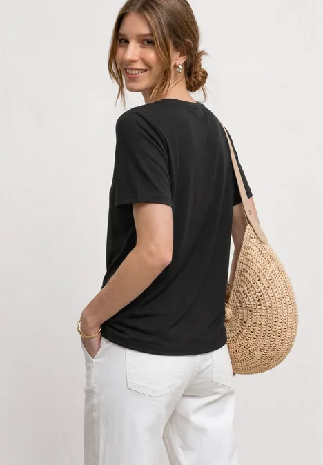 Frau in schwarzem T-Shirt und weißen Hosen mit geflochtener Stroh-Tasche, von hinten fotografiert.