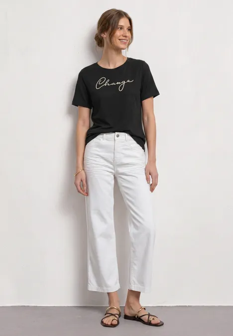 Eine Frau mit schwarzem T-Shirt, weißer Hose und flachen Sandalen vor hellem Hintergrund.