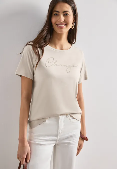 Vrouw draagt beige T-shirt met 'Challenge'-opdruk en witte broek, casual stijl.