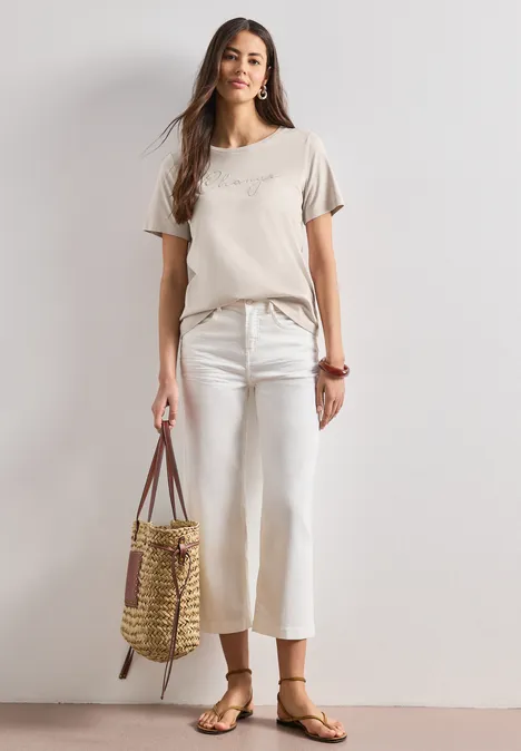 Vrouw in beige top, witte wijde broek en sandalen met een strooien tas voor lichte achtergrond