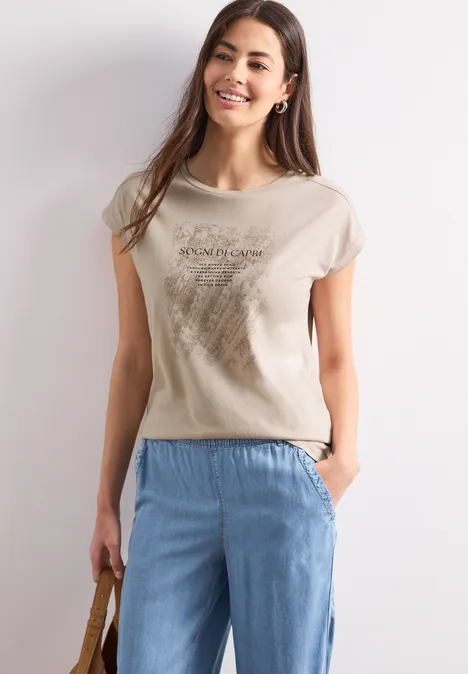 Een vrouw draagt een beige t-shirt en lichtblauwe broek, met een tas in haar hand.