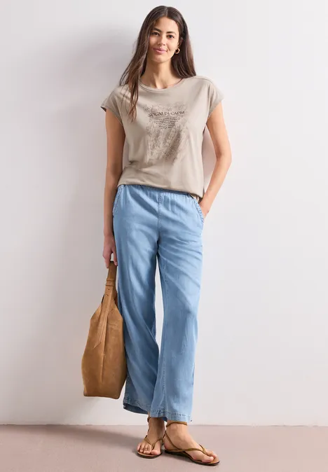 Vrouw in casual outfit: beige T-shirt, wijde blauwe jeans, sandalen en een handtas.