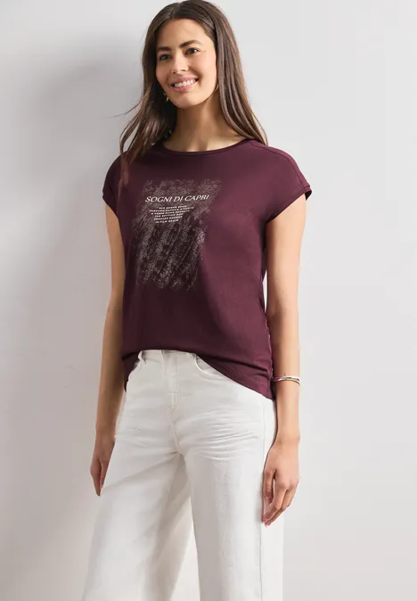 Frau mit bordeauxrotem T-Shirt und weißen Hosen vor hellem Hintergrund.