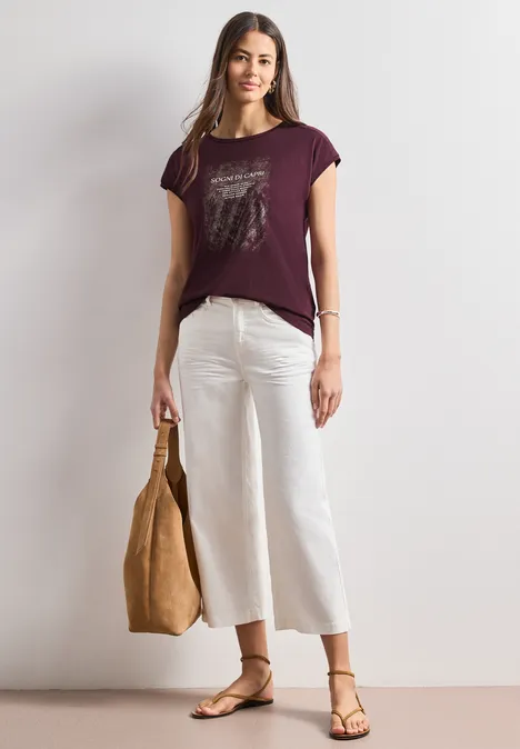 Frau mit unscharfem Gesicht trägt ein dunkelrotes T-Shirt, weiße Culottes, Sandalen und hält eine braune Tasche.