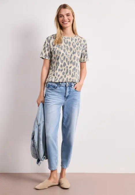 Femme en t-shirt à motifs, jean clair et mocassins, tenant une veste en jean.