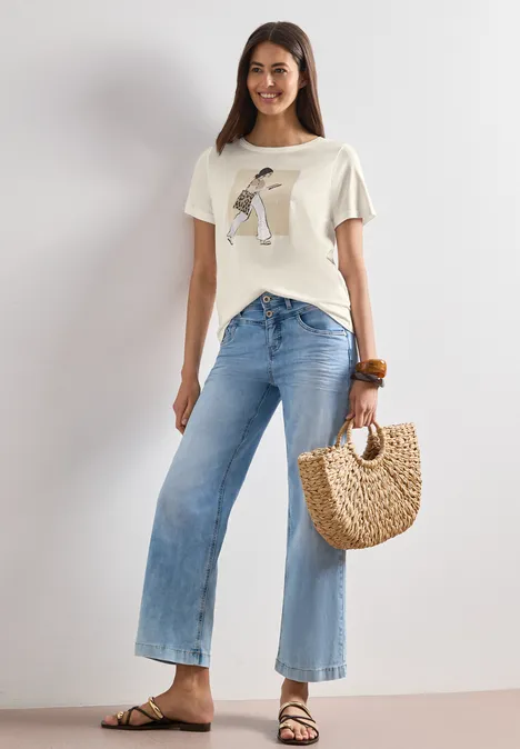 Frau trägt weißes T-Shirt mit Print, helle Schlagjeans, Basttasche und Sandalen.