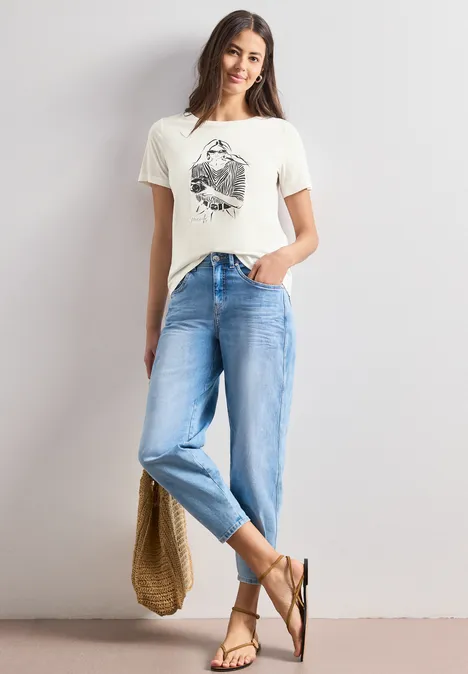 Eine Frau in beigem T-Shirt mit Print, blauen Jeans, Sandalen und Tasche steht vor weißer Wand.