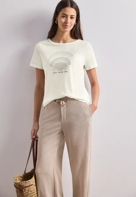 Frau in weißem T-Shirt mit Muster, beiger Hose und geflochtener Korbhandtasche.