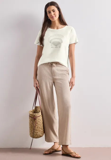 Frau in beigen Leinenhosen, weißem Print-T-Shirt, Strohtasche und Sandalen vor neutralem Hintergrund.