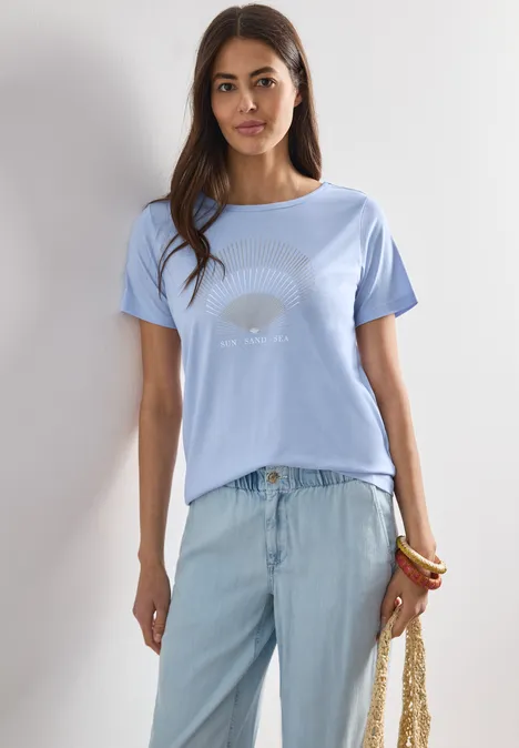 Frau trägt hellblaues T-Shirt, helle Jeans, hält geflochtene Tasche, trägt bunte Armbänder.