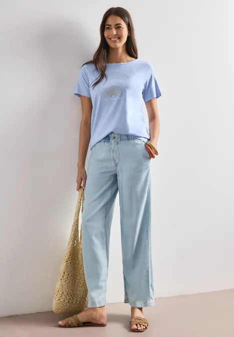 Frau in lockeren hellblauen Jeans, pastellblauem T-Shirt und großem Strohbeutel vor neutraler Wand.