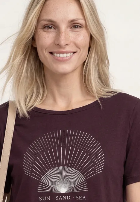 T-Shirt mit Rundhals und Foliendetails rustic rouge Frau mit hellblonden Haaren trägt ein dunkelviolettes Shirt mit Sonnendesign und dem Aufdruck 'Sun Sand Sea'.