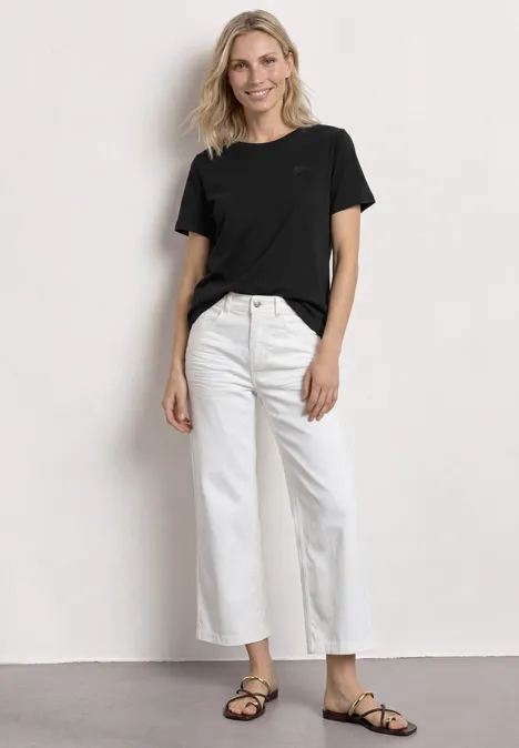 T-Shirt mit Rundhals und Embroidery Detail Black Frau mit schwarzem T-Shirt, weißen, weiten, knöchellangen Jeans und schwarzen Sandalen vor weißem Hintergrund.