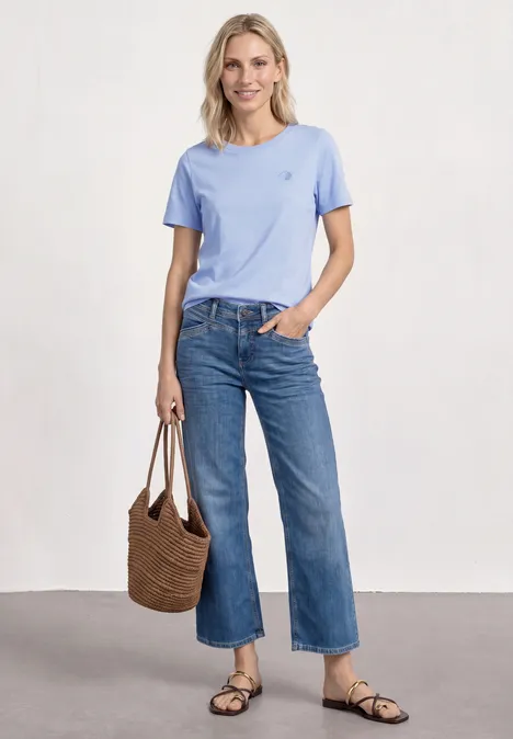 Vrouw met lichtblauw T-shirt, wijde cropped jeans, sandalen en rieten tas.