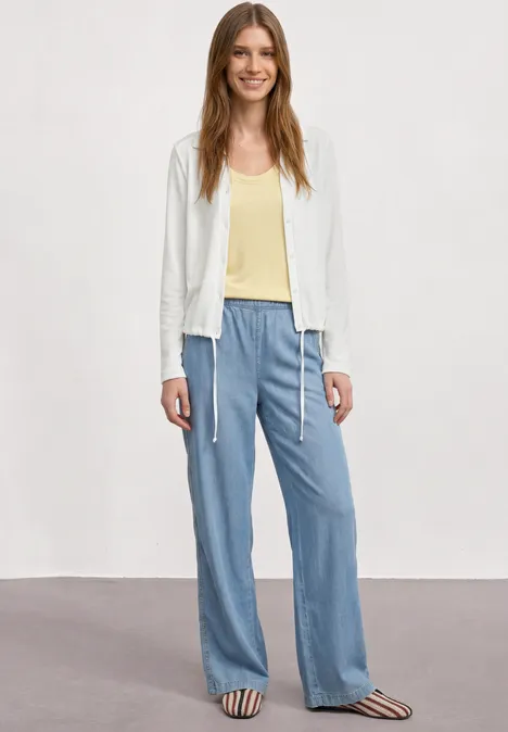 Leichte Langam Jacke in Leinen-Optik off white Eine Frau in weißer Jacke, gelbem Top, lockerer blauer Hose und gestreiften Sandalen vor schlichtem Hintergrund.