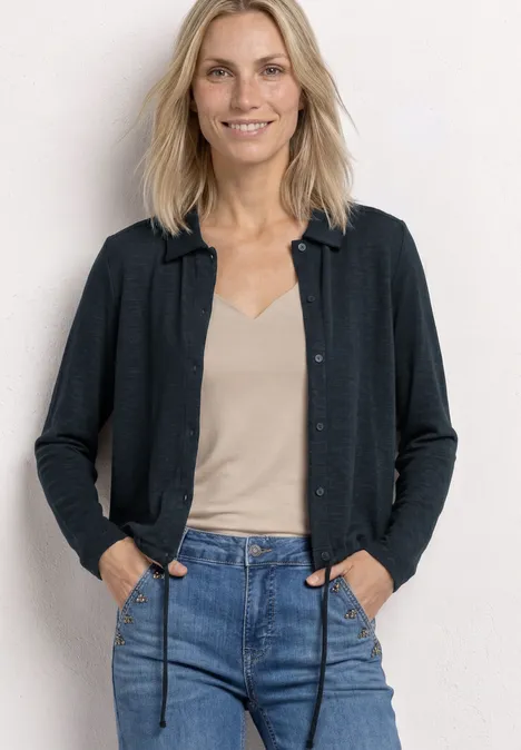Une femme aux cheveux blonds porte un jean, un débardeur beige et un cardigan gris devant un fond blanc.