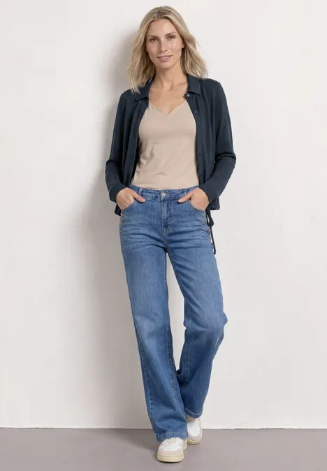 Femme devant un mur blanc, top beige, cardigan gris foncé, jean droit bleu et baskets blanches.